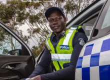 An man of African appearance sitting in a police car with the door open. He's wearing a police uniform, including hi-vis vest.