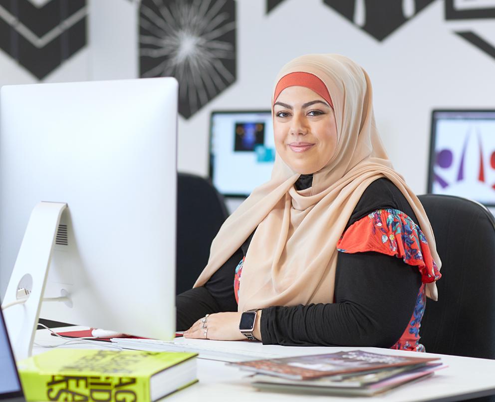 A woman in a hijab sitting at a mac computer with books and magazines nearby