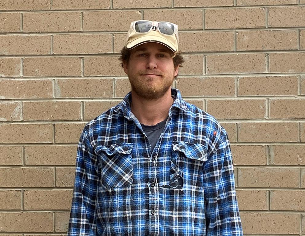 Tom Borg standing outside in front of a brick wall, wearing a cap and plaid shirt
